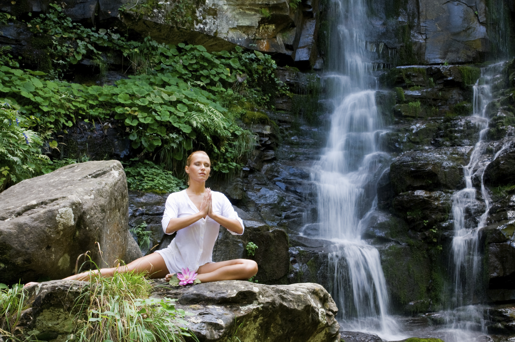 Beautiful young woman meditating with a lotus flower while doing yoga in a wonderful forest near waterfall